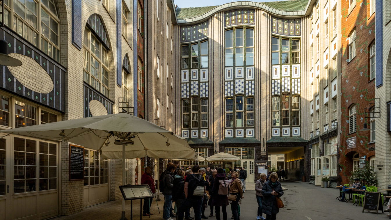 (c) Sonja Koller | 1000things Ein belebter Innenhof in den Hackeschen Höfen in Berlin mit kunstvoll gestalteten Jugendstilfassaden aus verzierten Fliesen und großen Fenstern. Mehrere Gruppen von Menschen stehen im Hof, einige unter großen Sonnenschirmen eines Restaurants mit Außenbereich. Die Architektur zeigt eine Mischung aus weißen, blauen und braunen Kacheln mit geometrischen Mustern. Auf der rechten Seite sind Cafés und Sitzbereiche mit grünen Stühlen zu sehen, während im Hintergrund ein Durchgang zu einem weiteren Hof führt.