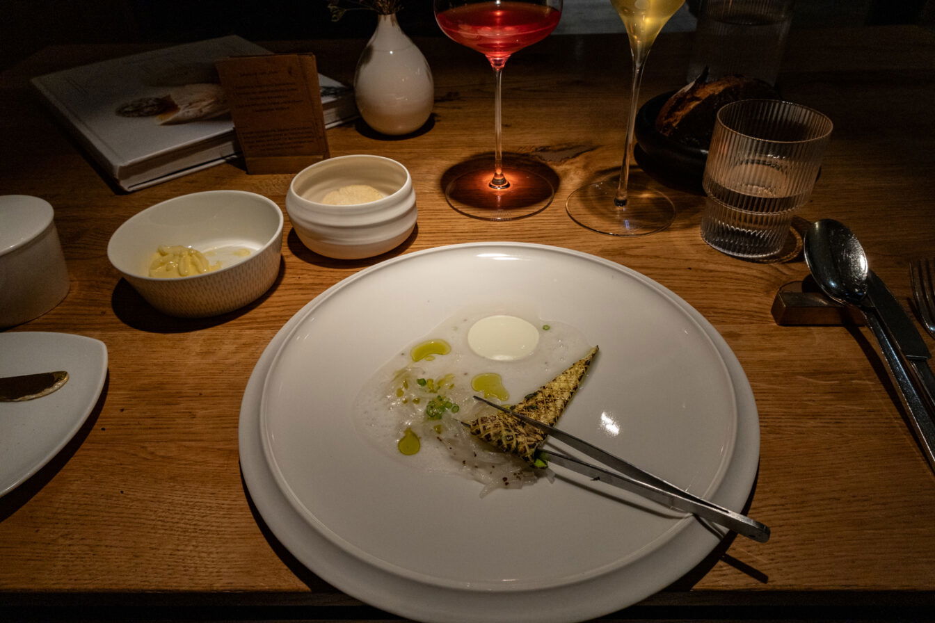 Ein kunstvoll angerichtetes Gericht im Fine-Dining-Restaurant Horváth in Berlin-Kreuzberg. Auf einem schlichten weißen Teller liegt eine filigrane Komposition aus dünn geschnittenem Fisch, einem luftigen Schaum, einem cremigen weißen Element sowie einem gegrillten Stück Gemüse, garniert mit feinen Kräutern und einigen Tropfen Öl. Eine Pinzette liegt auf dem Teller, was auf eine detailverliebte Präsentation hinweist.

Im Hintergrund auf dem elegant gedeckten Holztisch stehen feine Weingläser mit farbenfrohen Getränken, ein Wasserglas sowie kleine Schalen mit Butter und weiteren Beilagen. Ein dunkles Brot in einer schwarzen Schale deutet auf eine raffinierte Begleitung zum Menü hin. Die dezente, warme Beleuchtung verstärkt die intime Atmosphäre des Restaurants, das für seine moderne Interpretation der österreichischen Küche mit einem Fokus auf regionale Zutaten bekannt ist.