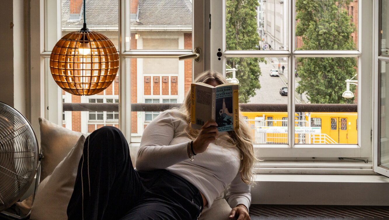 (c) Sonja Koller | 1000things Szene aus einem Zimmer des Michelberger Hotels in Berlin: Eine Frau liegt entspannt auf einer Holzplattform am Fenster, liest ein Buch und blickt dabei ins Freie. Hinter ihr ein großes, geöffnetes Sprossenfenster mit Blick auf eine ruhige Berliner Straße, hohe Bäume und eine vorbeifahrende gelbe Straßenbahn. Links im Bild eine hängende, kugelförmige Lampe aus Holzstreben und ein Ventilator auf einem Kissen. Die Atmosphäre ist gemütlich, urban und entspannt.