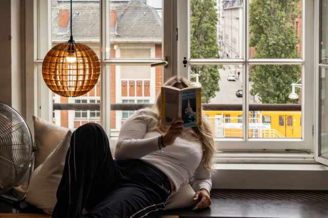 Szene aus einem Zimmer des Michelberger Hotels in Berlin: Eine Frau liegt entspannt auf einer Holzplattform am Fenster, liest ein Buch und blickt dabei ins Freie. Hinter ihr ein großes, geöffnetes Sprossenfenster mit Blick auf eine ruhige Berliner Straße, hohe Bäume und eine vorbeifahrende gelbe Straßenbahn. Links im Bild eine hängende, kugelförmige Lampe aus Holzstreben und ein Ventilator auf einem Kissen. Die Atmosphäre ist gemütlich, urban und entspannt.