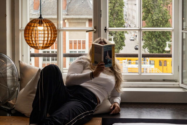 Szene aus einem Zimmer des Michelberger Hotels in Berlin: Eine Frau liegt entspannt auf einer Holzplattform am Fenster, liest ein Buch und blickt dabei ins Freie. Hinter ihr ein großes, geöffnetes Sprossenfenster mit Blick auf eine ruhige Berliner Straße, hohe Bäume und eine vorbeifahrende gelbe Straßenbahn. Links im Bild eine hängende, kugelförmige Lampe aus Holzstreben und ein Ventilator auf einem Kissen. Die Atmosphäre ist gemütlich, urban und entspannt.