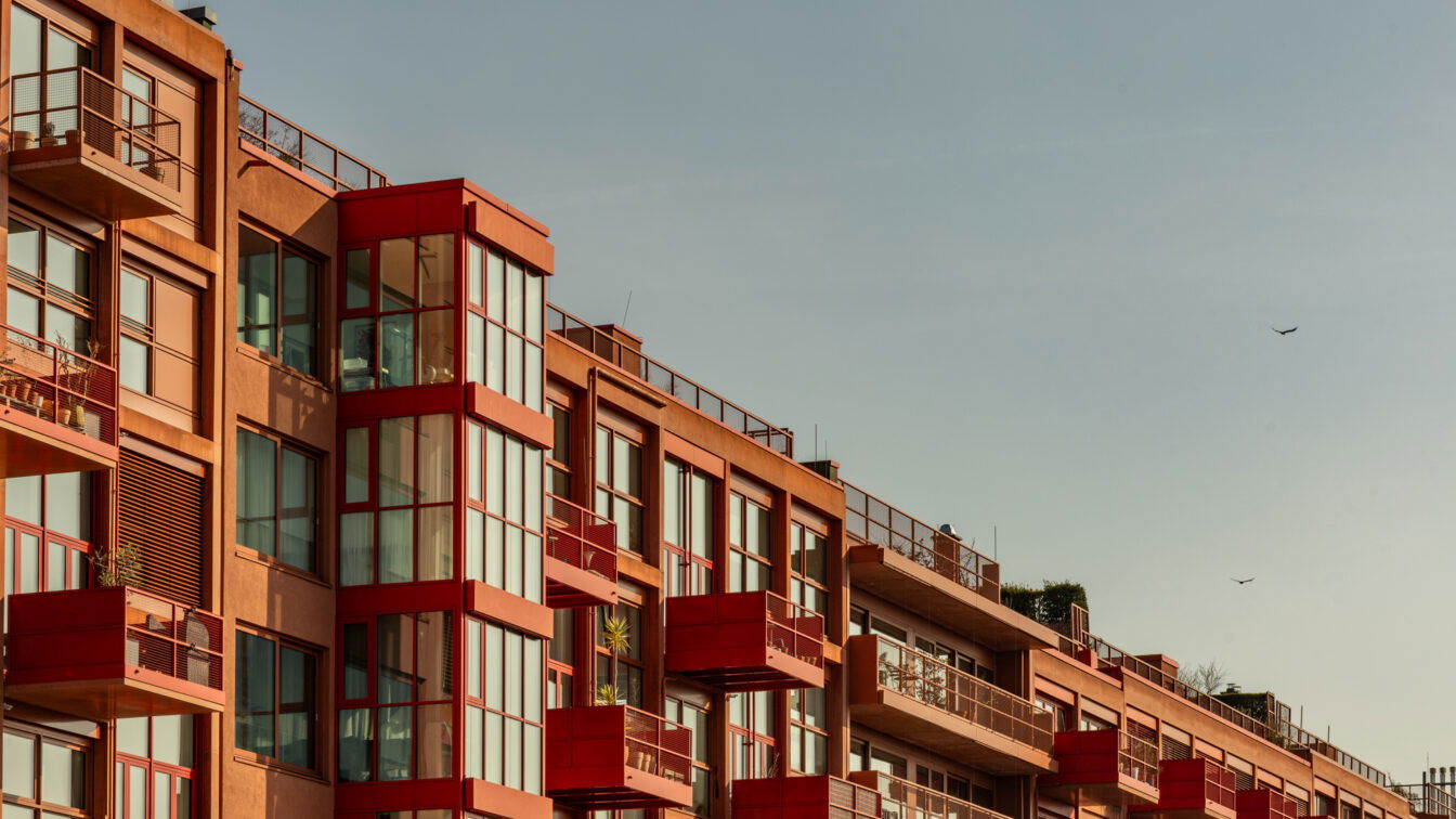 Eine moderne Wohnhausfassade in Berlin-Kreuzberg mit markanten roten Balkonen und großzügigen Glasfronten, die das Sonnenlicht reflektieren. Die Architektur kombiniert klare Linien mit warmen Erdtönen, während die roten Akzente einen auffälligen Kontrast setzen. Die Balkone sind teilweise mit Pflanzen dekoriert, was die urbane, aber dennoch wohnliche Atmosphäre unterstreicht. Im oberen Bereich sind Dachterrassen mit Geländern zu sehen, die einen offenen Blick über die Stadt ermöglichen. Der Himmel ist weit und klar, mit wenigen Vögeln, die in der Ferne fliegen
