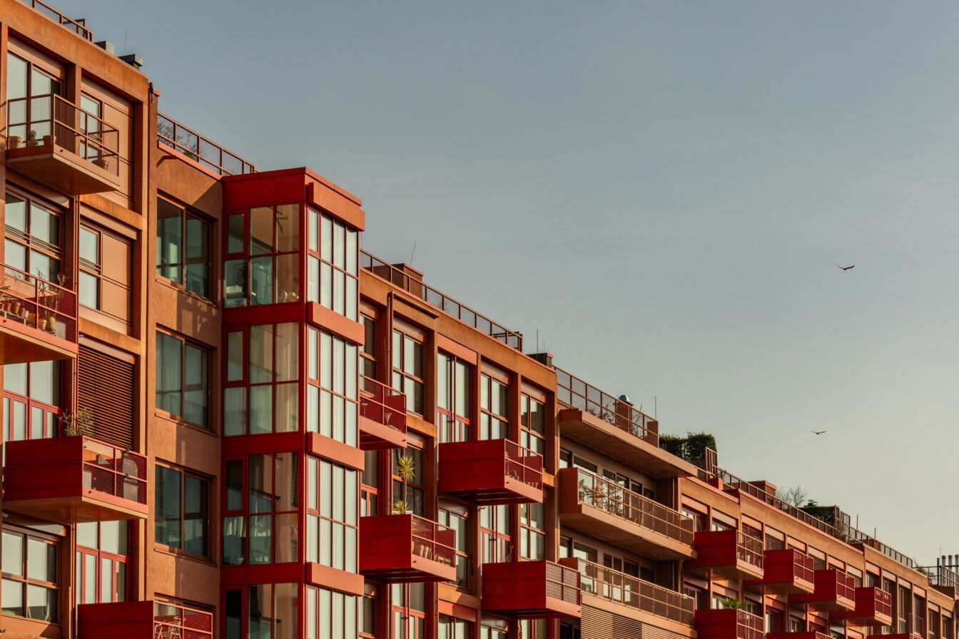 Eine moderne Wohnhausfassade in Berlin-Kreuzberg mit markanten roten Balkonen und großzügigen Glasfronten, die das Sonnenlicht reflektieren. Die Architektur kombiniert klare Linien mit warmen Erdtönen, während die roten Akzente einen auffälligen Kontrast setzen. Die Balkone sind teilweise mit Pflanzen dekoriert, was die urbane, aber dennoch wohnliche Atmosphäre unterstreicht. Im oberen Bereich sind Dachterrassen mit Geländern zu sehen, die einen offenen Blick über die Stadt ermöglichen. Der Himmel ist weit und klar, mit wenigen Vögeln, die in der Ferne fliegen