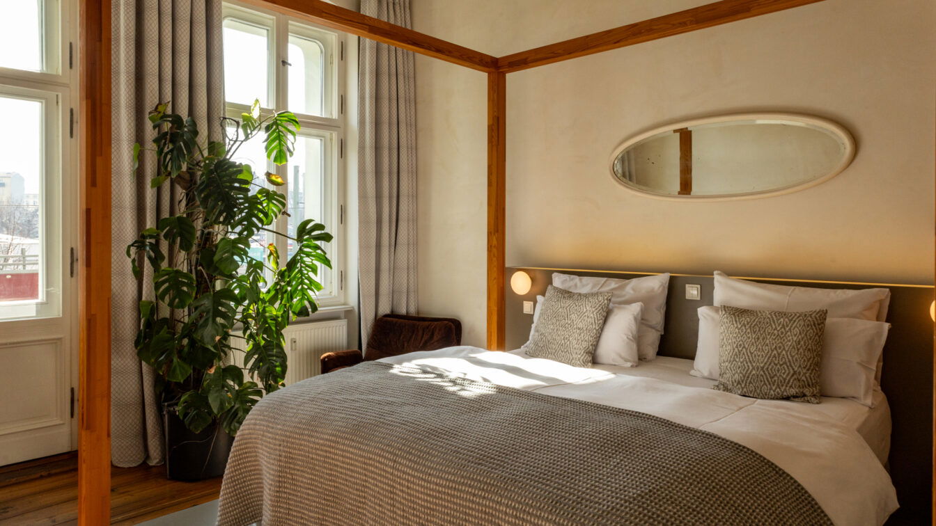 (c) Sonja Koller | 1000things Ein stilvoll eingerichtetes Hotelzimmer mit einem gemütlichen Doppelbett, das mit weißen Kissen und einer grau gemusterten Decke dekoriert ist. Das Bett hat ein modernes, minimalistisches Kopfteil mit integrierten Wandlampen. Über dem Bett hängt ein ovaler Spiegel. Das Zimmer ist von natürlichem Licht durchflutet, das durch große Fenster mit hellgrauen Vorhängen fällt. Eine große grüne Pflanze steht in der Ecke, daneben ein brauner Sessel. Die Decke zeigt dezente historische Stuckelemente, und der Holzboden verleiht dem Raum eine warme Atmosphäre.