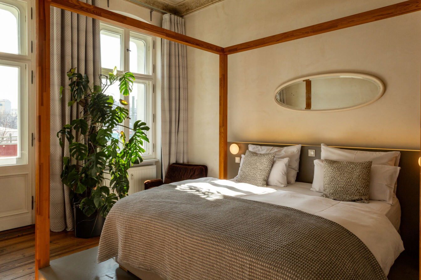 Ein stilvoll eingerichtetes Hotelzimmer mit einem gemütlichen Doppelbett, das mit weißen Kissen und einer grau gemusterten Decke dekoriert ist. Das Bett hat ein modernes, minimalistisches Kopfteil mit integrierten Wandlampen. Über dem Bett hängt ein ovaler Spiegel. Das Zimmer ist von natürlichem Licht durchflutet, das durch große Fenster mit hellgrauen Vorhängen fällt. Eine große grüne Pflanze steht in der Ecke, daneben ein brauner Sessel. Die Decke zeigt dezente historische Stuckelemente, und der Holzboden verleiht dem Raum eine warme Atmosphäre.