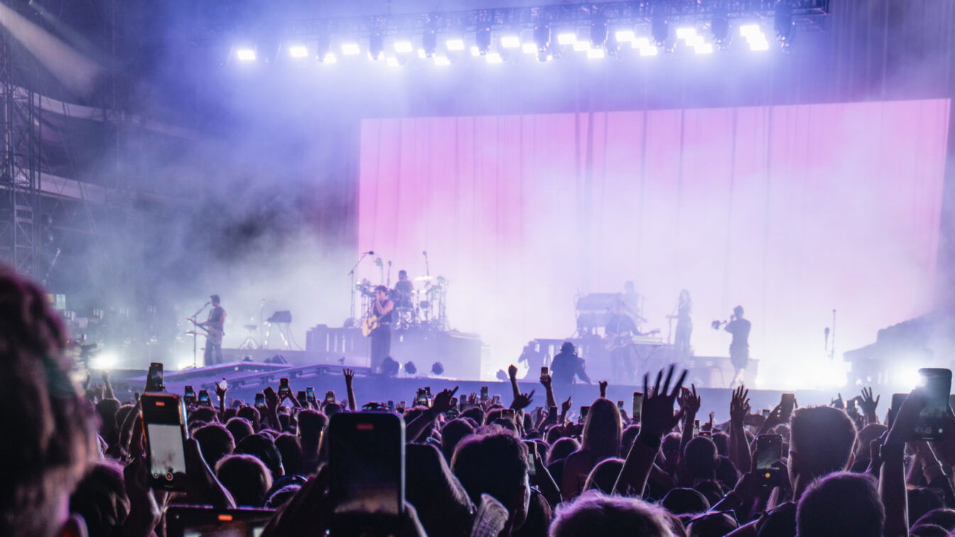 Große Menschenmenge bei einem Open-Air-Festival in Europa, viele halten ihre Smartphones hoch. Auf der Bühne spielen mehrere Musiker vor einer großen, rosa beleuchteten Leinwand, umgeben von Nebel und starken Scheinwerfern.