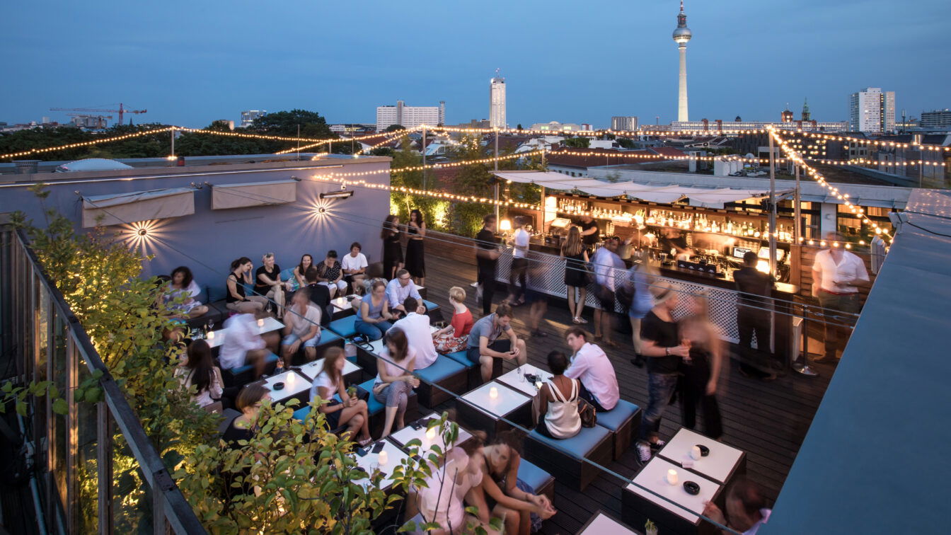 Dachterrasse mir Bar und Tischen an denen Personen sitzen. Im Hintergrund sieht man Berlin und den Fernsehturm