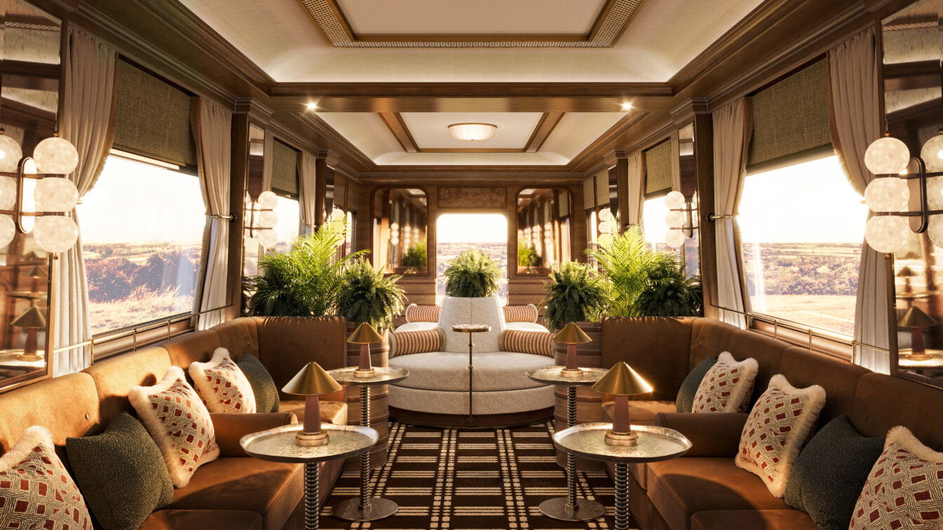 (c) Belmond Luxuriöses Interieur eines historischen Zugwaggons mit edler Holzverkleidung, großen Fenstern und stilvoller Beleuchtung. Die Lounge ist mit samtigen Polstern in warmen Brauntönen, dekorativen Kissen und eleganten Beistelltischen mit goldenen Lampen ausgestattet. Üppige grüne Pflanzen verleihen dem Raum eine frische Note, während ein geometrisch gemusterter Teppich den Boden schmückt. Die Fenster geben den Blick auf eine malerische Landschaft frei, die in warmes Sonnenlicht getaucht ist.