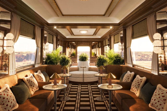Luxuriöses Interieur eines historischen Zugwaggons mit edler Holzverkleidung, großen Fenstern und stilvoller Beleuchtung. Die Lounge ist mit samtigen Polstern in warmen Brauntönen, dekorativen Kissen und eleganten Beistelltischen mit goldenen Lampen ausgestattet. Üppige grüne Pflanzen verleihen dem Raum eine frische Note, während ein geometrisch gemusterter Teppich den Boden schmückt. Die Fenster geben den Blick auf eine malerische Landschaft frei, die in warmes Sonnenlicht getaucht ist.