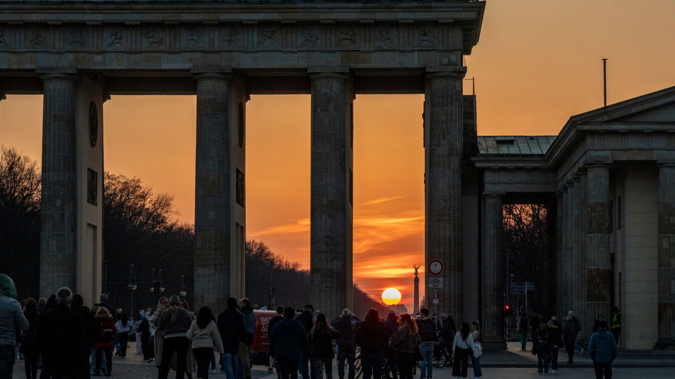 Menschen spazieren bei Sonnenuntergang durch das Brandenburger Tor in Berlin. Die Sonne steht tief am Horizont und scheint genau durch die Säulen des Tores. Der Himmel ist orange gefärbt, Silhouetten der Menschen und Gebäude rahmen die Szene stimmungsvoll ein.