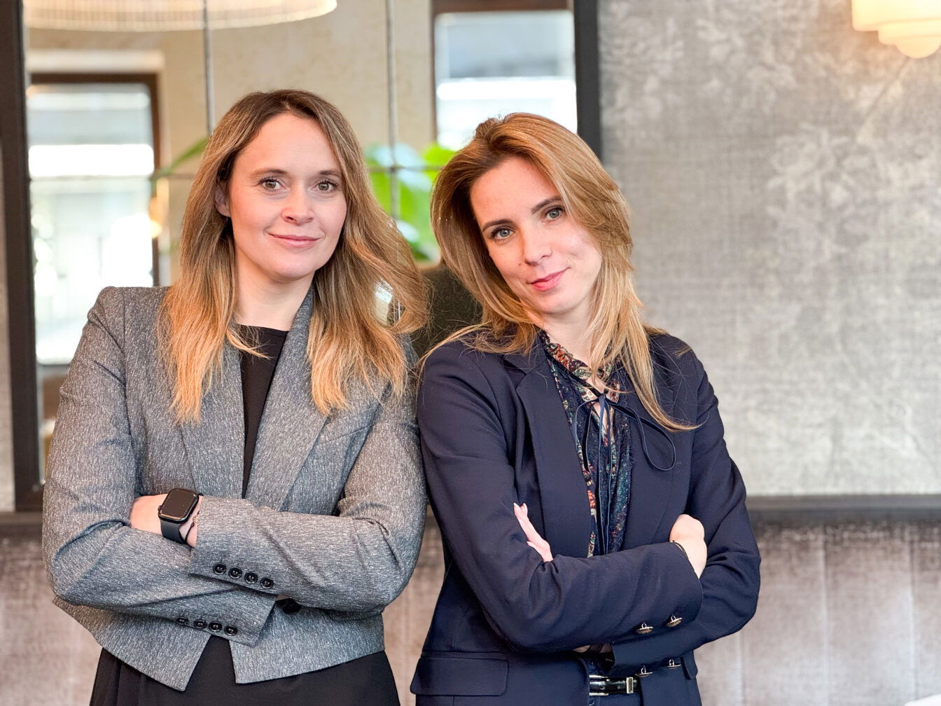 Zwei Frauen in Business-Outfits stehen mit verschränkten Armen nebeneinander in einem modernen Innenraum mit Spiegeln und stilvoller Beleuchtung. Beide lächeln selbstbewusst in die Kamera