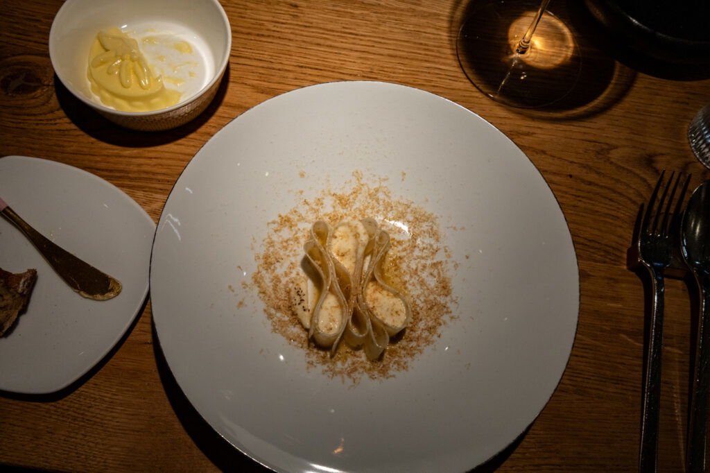Ein kunstvoll angerichtetes Pastagericht auf einem weißen Teller: Die Pasta ist elegant gefaltet und mit einer cremigen Sauce überzogen, umgeben von feinen, geriebenen Zutaten. Die Szene ist in warmem Licht gehalten, mit einem Glas Wein und weiteren Begleitkomponenten auf einem rustikalen Holztisch.