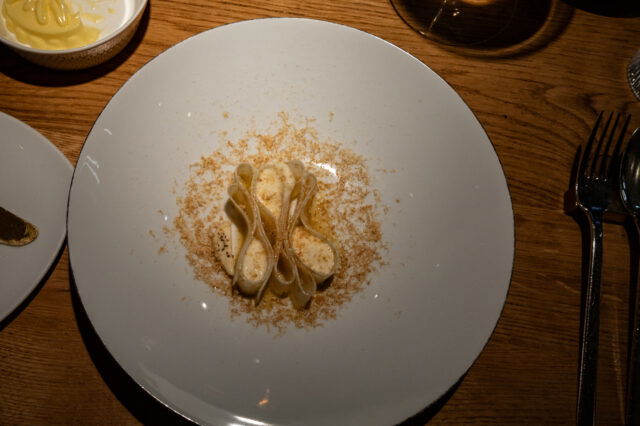 Ein kunstvoll angerichtetes Pastagericht auf einem weißen Teller: Die Pasta ist elegant gefaltet und mit einer cremigen Sauce überzogen, umgeben von feinen, geriebenen Zutaten. Die Szene ist in warmem Licht gehalten, mit einem Glas Wein und weiteren Begleitkomponenten auf einem rustikalen Holztisch.