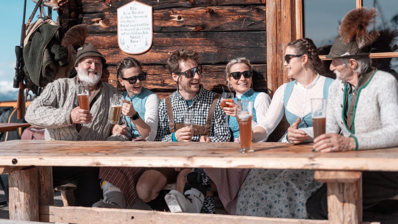 Personen in Tracht sitzen vor der Hütte und stoßen gutgelaunt an