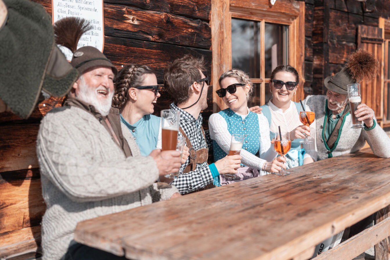 Personen in Tracht sitzen vor der Hütte und stoßen gutgelaunt an