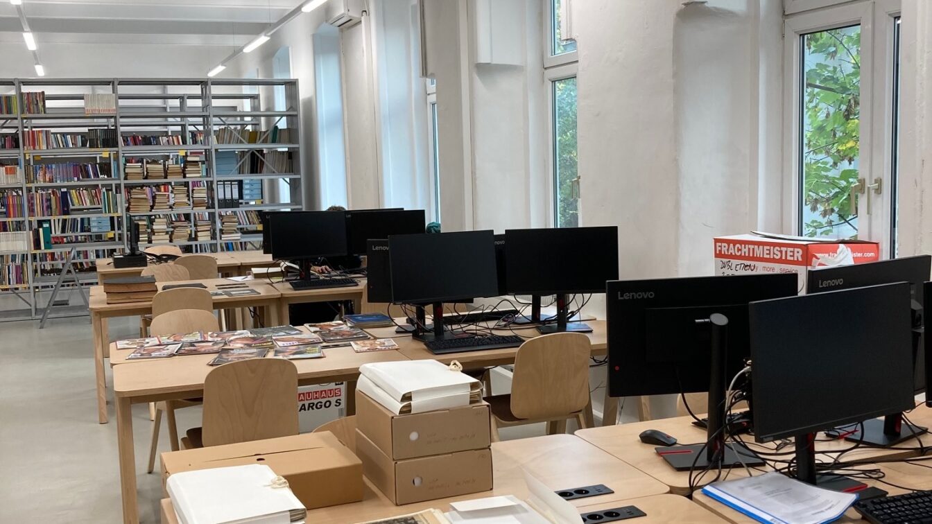 (c) Hannes Sulzenbacher | QWIEN Das Bild zeigt einen modernen Raum, der wie ein Arbeitsbereich in einer Bibliothek oder einem Archiv aussieht. Es gibt mehrere Tische mit Computern und Büchern. Auf den Tischen liegen Zeitschriften und einige Dokumente, die teilweise in weiße Ordner verpackt sind. Im Hintergrund sind Regale mit einer Vielzahl von Büchern sichtbar. Große Fenster lassen natürliches Licht in den Raum, und im hinteren Bereich des Raums ist ein Kasten mit Aufschrift "Frachtmeister" zu sehen. Ein Mitarbeiter ist an einem der Computer zu sehen, während er an einem Bildschirm arbeitet.