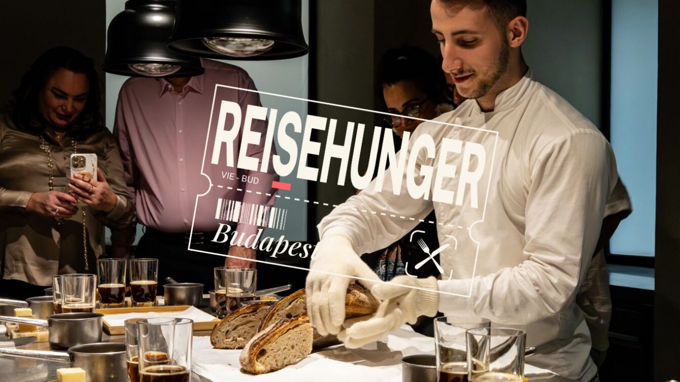 (c) Sonja Koller | 1000things Ein junger Koch in weißer Kochjacke und Handschuhen schneidet frisches Sauerteigbrot bei einem Tasting im Onyx Műhely in Budapest. Um ihn herum stehen mehrere Gäste, die das Erlebnis fotografieren und beobachten. Auf dem Tisch befinden sich Gläser mit Getränken und kleine Töpfe. Im Vordergrund ist der Schriftzug „REISEHUNGER – VIE-BUD – Budapest“ eingeblendet.