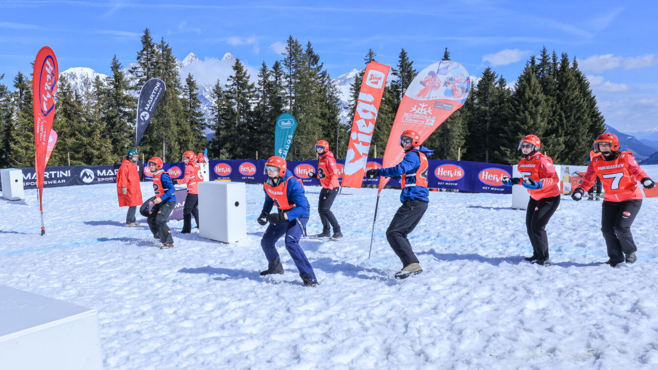 (c) Filzmoos Tourismus | Michael Simonlehner Eine Gruppe von Menschen nimmt an einer Schneeballschlacht-Wettbewerb teil. Sie tragen Helme, Schutzbrillen und farbige Trikots mit Nummern. Die Szene spielt sich auf einer schneebedeckten Fläche in den Bergen ab, mit blauen Bannern und bunten Fahnen im Hintergrund. Die Teilnehmer bewegen sich aktiv, einige in Abwehrhaltung, andere scheinbar zum Wurf bereit. Die Sonne scheint, und der Himmel ist klar mit wenigen Wolken. Im Hintergrund sind Nadelbäume und schneebedeckte Berggipfel zu sehen.