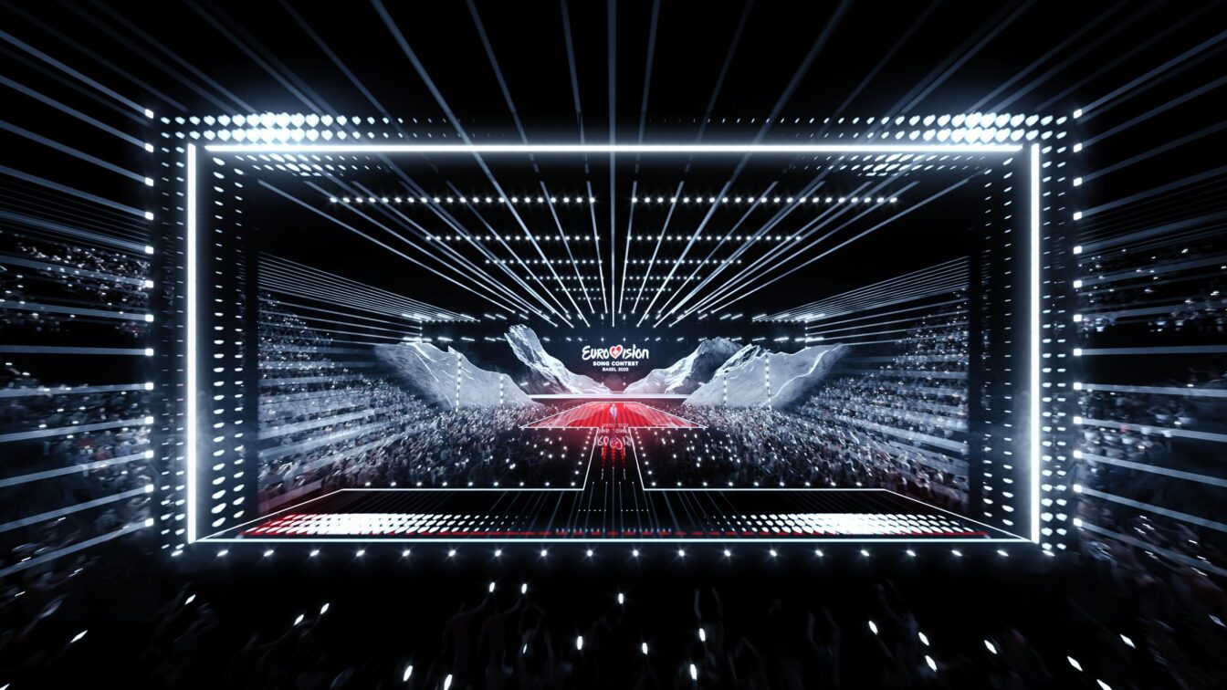 (c) Songcontest 2025 Das Bild zeigt eine beeindruckende Szene vom Eurovision Song Contest 2025 in Basel, mit einer modernen und futuristischen Bühne. Die Bühne ist von hellen Lichtstrahlen umgeben, die dynamisch durch den Raum verlaufen und den Fokus auf das große Event lenken. Auf der Bühne ist das "Eurovision Song Contest"-Logo prominent zu sehen. Im Hintergrund ist das Publikum mit vielen Handylichtern zu sehen, die eine festliche Atmosphäre schaffen. Die Beleuchtung und das Design der Bühne unterstreichen das spektakuläre und energiegeladene Ambiente dieses internationalen Musikwettbewerbs.
