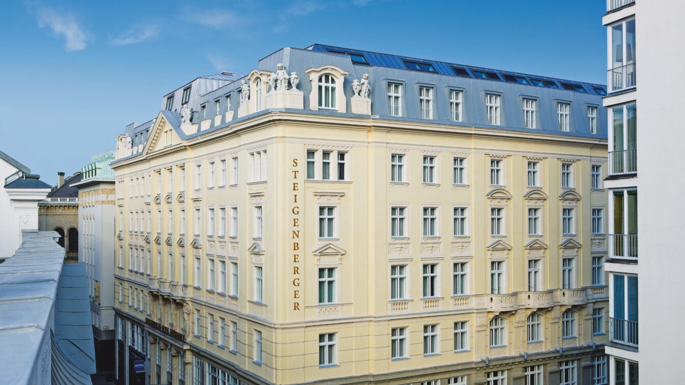 (c) Steigenberger Hotel Außenansicht des Steigenberger Hotels, ein elegantes, hellgelbes Gebäude mit klassischer Architektur, zahlreichen Fenstern und kunstvollen Fassadendetails. Der Hotelname ist vertikal an der Seite der Fassade zu sehen, der Himmel ist blau und klar.