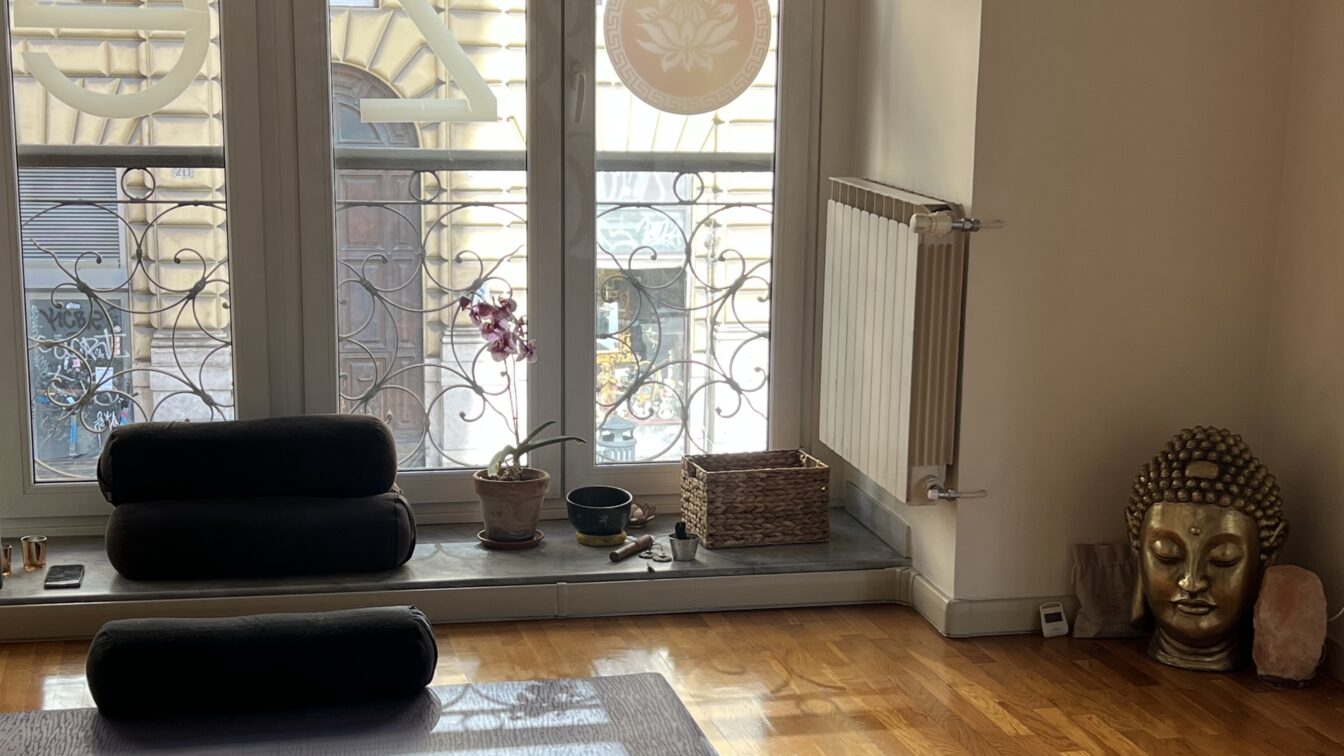 (c) Una Arslanagic Dieses Bild zeigt einen einladenden Yoga- oder Meditationsraum mit einer warmen und beruhigenden Atmosphäre. Im Vordergrund liegt eine Yogamatte mit einem schwarzen Bolster darauf. Weitere Bolster sind auf der Fensterbank gestapelt. Auf der Fensterbank befinden sich zudem eine Orchidee, eine kleine Schale und ein geflochtener Korb mit verschiedenen Gegenständen. Rechts im Raum steht eine große goldene Buddha-Kopf-Skulptur neben einem Salzkristalllicht. Durch die großen Fenster mit kunstvollen Gitterverzierungen fällt natürliches Licht ein, und im Hintergrund ist eine städtische Umgebung mit verzierten Gebäudefassaden zu sehen.