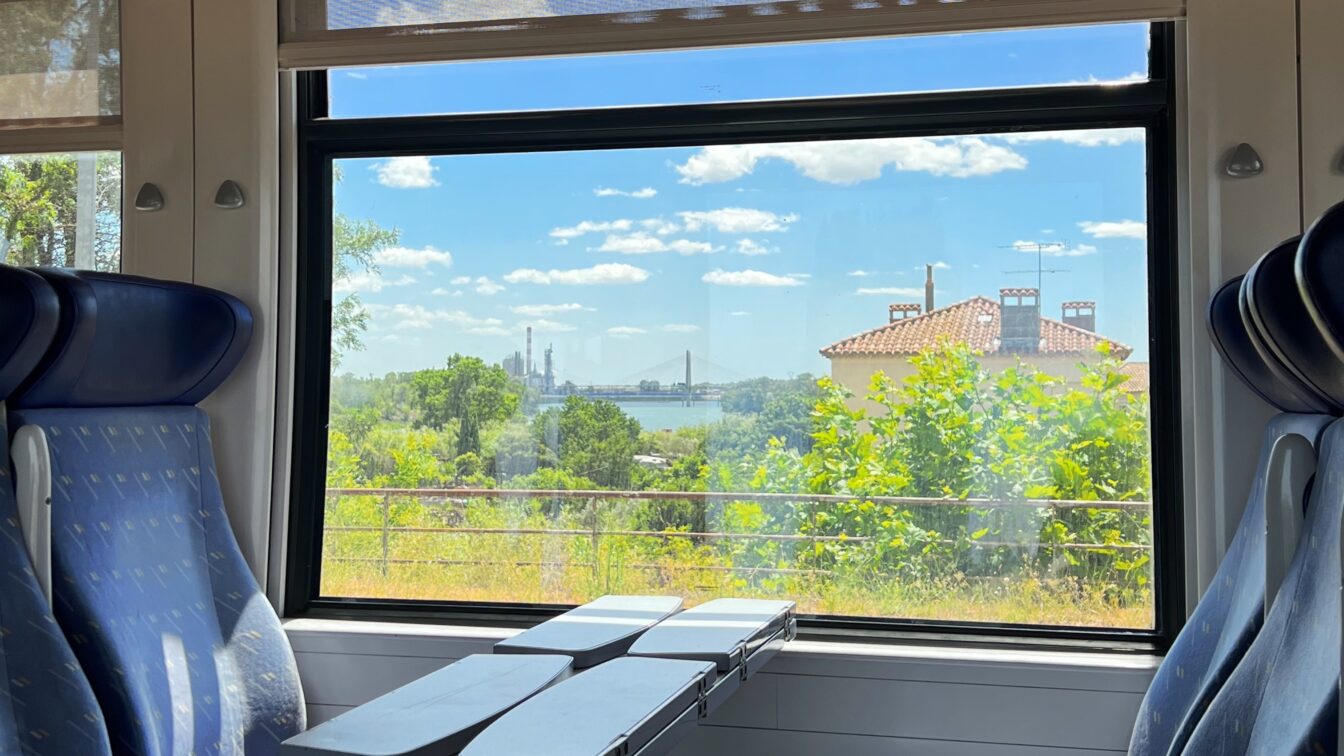 (c) Grace McCuistion | dupe Zugfahrt Frankreich Barcelona Italien, Blick aus dem Fenster eines Zugabteils mit blauen Sitzen und Klapptischen. Draußen ist eine grüne Landschaft mit Bäumen, einem Haus mit rotem Ziegeldach und einer Brücke im Hintergrund zu sehen. Der Himmel ist blau mit einigen weißen Wolken.