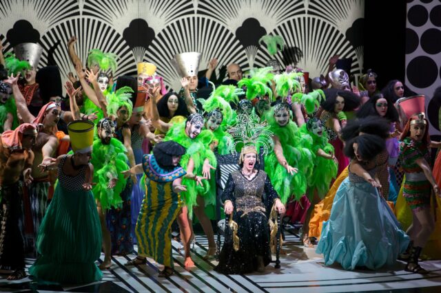 Schillernde Bühnensituation mit vielen Personen in bunten Kostümen mit grünen Federn und auffällig geschminkten Gesichtern