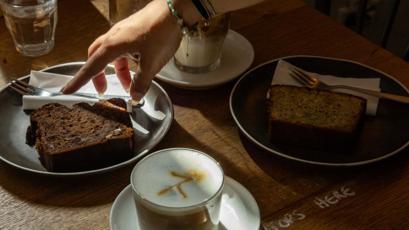 (c) Sonja Koller | 1000things Gemütliche Café-Szene in Berlin: Auf einem Holztisch stehen zwei Stücke Kuchen auf dunklen Tellern, Cappuccino mit Latte Art und ein Glas Wasser. Eine Hand greift nach Besteck im warmen Licht – typisches Berliner Café-Flair mit entspannter Atmosphäre und hausgemachtem Gebäck.