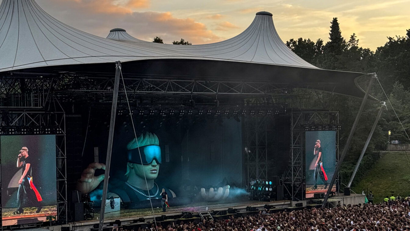 Großes Open-Air-Konzert im Sommer 2025 in Berlin: Eine riesige Menschenmenge steht vor der überdachten Bühne der Waldbühne. Auf der Bühne ein Künstler mit Sonnenbrille, unterstützt von beeindruckenden LED-Effekten und einer riesigen futuristischen Bühnenfigur. Der Himmel zeigt ein farbenfrohes Abendlicht – perfekte Festivalstimmung in Berlin.