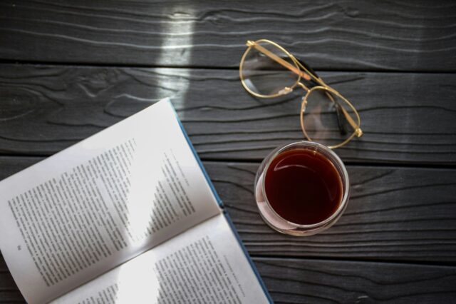 Ein aufgeschlagenes Buch neben einem Glas Rotwein und einer Lesebrille.