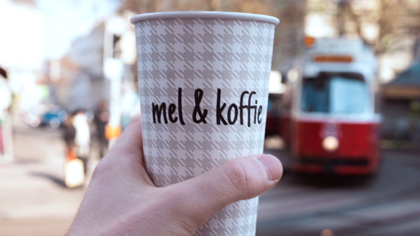 (c) mel&koffie Ein Kaffeebecher von Mel&koffie, im Hintergrund ist verschwommen eine rote Wiener Straßenbahn zu sehen