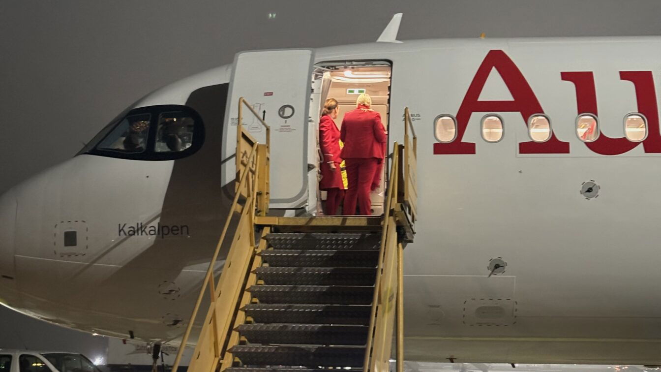 (c) Sonja Koller | 1000things Ein Flugzeug der Austrian Airlines steht bei Nacht auf dem Rollfeld des Flughafens. Drei Crewmitglieder in roten Uniformen stehen in der geöffneten Flugzeugtür und unterhalten sich. Eine mobile Treppe führt zur Tür hinauf. Der Flugzeugname „Kalkalpen“ ist neben dem Cockpit sichtbar, wo zwei Pilot*innen sitzen. Die Szene ist leicht nebelig beleuchtet.