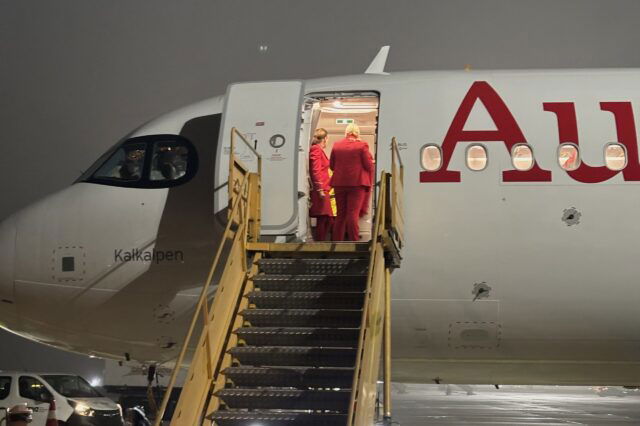 Ein Flugzeug der Austrian Airlines steht bei Nacht auf dem Rollfeld des Flughafens. Drei Crewmitglieder in roten Uniformen stehen in der geöffneten Flugzeugtür und unterhalten sich. Eine mobile Treppe führt zur Tür hinauf. Der Flugzeugname „Kalkalpen“ ist neben dem Cockpit sichtbar, wo zwei Pilot*innen sitzen. Die Szene ist leicht nebelig beleuchtet.