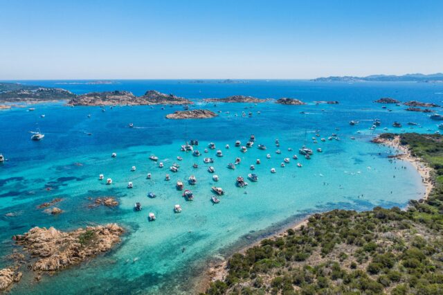 Luftaufnahme vom türkisblauen Meer vor Sardinien mit zahlreichen Booten vor der Küste und feinem Sandstrand – beliebtes Sommerziel in Italien mit kristallklarem Wasser und mediterraner Landschaft.
