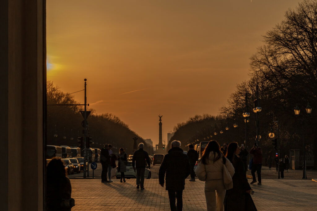 Menschen spazieren bei Sonnenuntergang auf der Straße des 17. Juni in Berlin, mit Blick auf die goldene Siegessäule in der Ferne. Die tiefstehende Sonne taucht den Himmel in warme Orangetöne, während Laternenlichter entlang der Straße leuchten. Bäume säumen den Weg, Silhouetten und Schatten betonen die ruhige Abendstimmung.