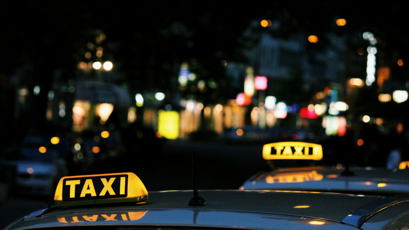 Zwei beleuchtete Taxi-Schilder auf den Dächern weißer Autos in einer nächtlichen, unscharf beleuchteten Stadtstraße. Im Hintergrund leuchten verschwommene Lichtquellen von Geschäften, Autos und Straßenlaternen.