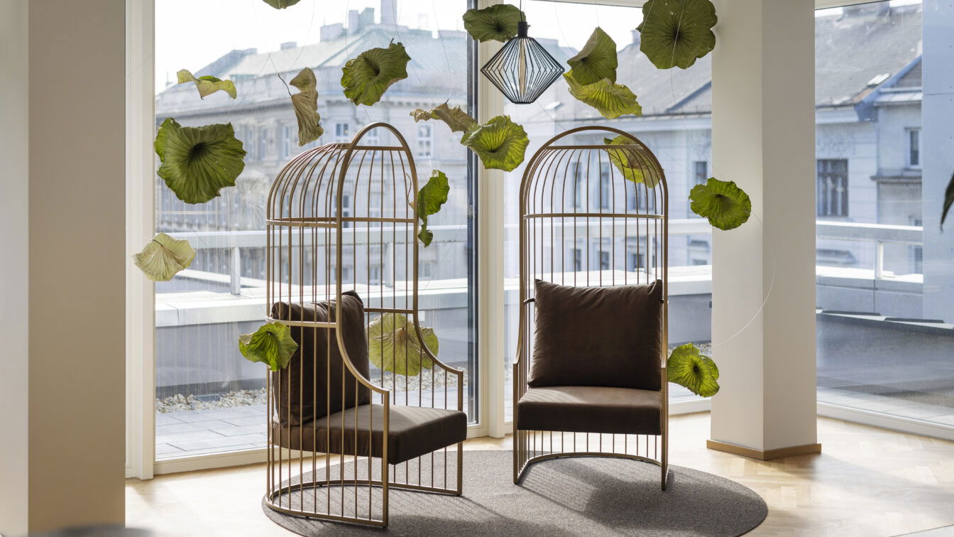 (c) ARCOTEL Wien Zwei moderne, vogelkäfigartige Sessel mit braunen Polstern stehen einander gegenüber auf einem runden Teppich in einem lichtdurchfluteten Raum mit Panoramafenstern im ARCOTEL Wien. Zwischen den Sesseln schweben dekorative grüne Blätter, die an dünnen Fäden von der Decke hängen. Im Hintergrund ist die Wiener Innenstadt zu sehen