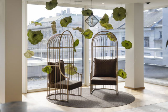 Zwei moderne, vogelkäfigartige Sessel mit braunen Polstern stehen einander gegenüber auf einem runden Teppich in einem lichtdurchfluteten Raum mit Panoramafenstern im ARCOTEL Wien. Zwischen den Sesseln schweben dekorative grüne Blätter, die an dünnen Fäden von der Decke hängen. Im Hintergrund ist die Wiener Innenstadt zu sehen