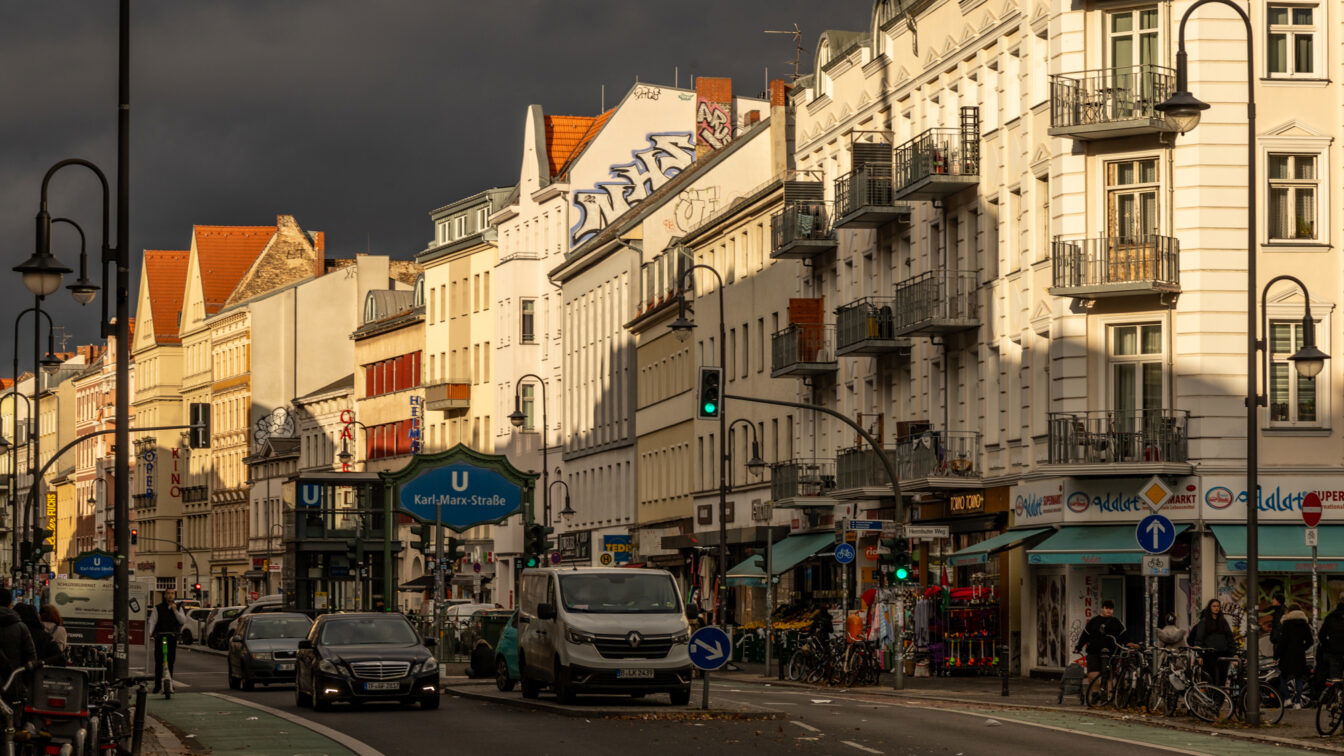 (c) Sonja Koller | 1000things Straßenszene aus Berlin-Neukölln bei dramatischem Licht. Die untergehende Sonne beleuchtet eine Häuserzeile mit Altbaufassaden, während dunkle Wolken den Himmel verdunkeln. Im Vordergrund ist der U-Bahnhof „Karl-Marx-Straße“ zu sehen, darüber prangt ein großes Graffiti. Autos, Fahrräder und Passant*innen beleben die Straße. Läden und ein Kino sind deutlich sichtbar.
