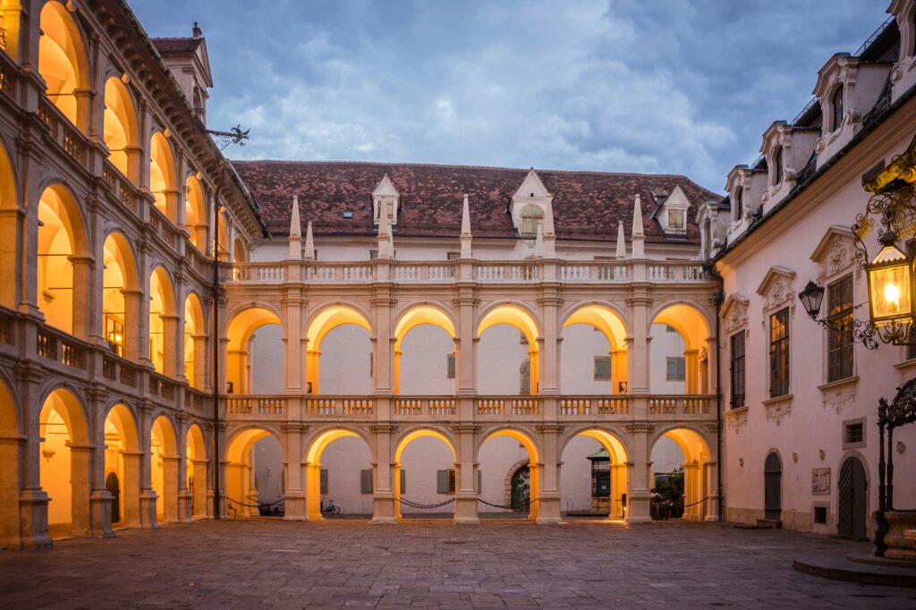 Innenhof der Grazer Burg im Abendlicht mit zwei Stockwerken offener Arkadengänge im Renaissancestil. Die warm beleuchteten Rundbögen und das Kopfsteinpflaster erzeugen eine stimmungsvolle Atmosphäre. Über dem Hof wölbt sich ein bewölkter Abendhimmel.