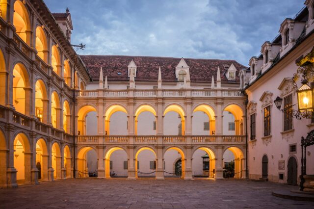 Innenhof der Grazer Burg im Abendlicht mit zwei Stockwerken offener Arkadengänge im Renaissancestil. Die warm beleuchteten Rundbögen und das Kopfsteinpflaster erzeugen eine stimmungsvolle Atmosphäre. Über dem Hof wölbt sich ein bewölkter Abendhimmel.