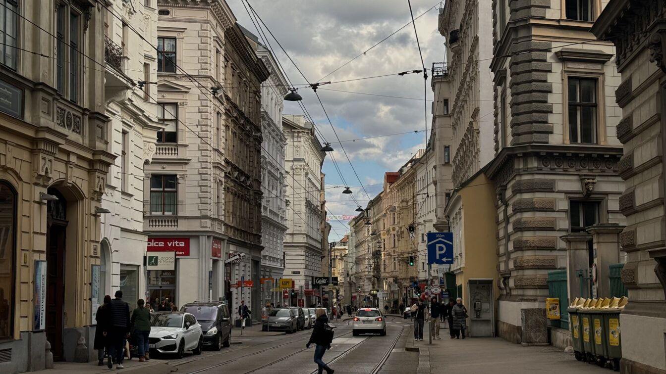 (c) 1000things Straßenszene auf der Josefstäder Straße in Wien, Blickrichtung stadteinwärts. Historische Fassaden säumen die Straße, an der Straßenbahnschienen verlaufen. Menschen gehen am Gehsteig, Autos parken am Rand, ein paar fahren in der Mitte. Bewölkter Himmel mit Sonnenstrahlen, die durch die Wolken brechen.