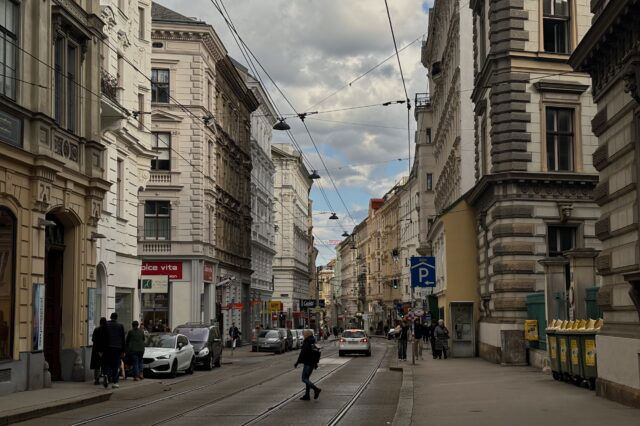 (c) 1000things Straßenszene auf der Josefstäder Straße in Wien, Blickrichtung stadteinwärts. Historische Fassaden säumen die Straße, an der Straßenbahnschienen verlaufen. Menschen gehen am Gehsteig, Autos parken am Rand, ein paar fahren in der Mitte. Bewölkter Himmel mit Sonnenstrahlen, die durch die Wolken brechen.