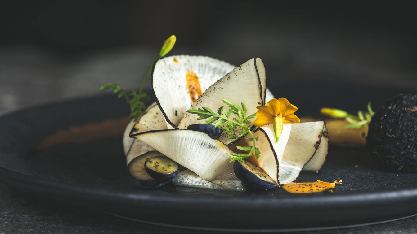 (c) Mario Stockhausen Gourmetgericht im Restaurant Kräuterreich by Vitus Winkler: kunstvoll angerichtete Komposition aus hauchdünnem Gemüse, Wildkräutern, Blüten und feinen Texturen auf dunklem Teller – moderne Alpenküche auf Sterne-Niveau.