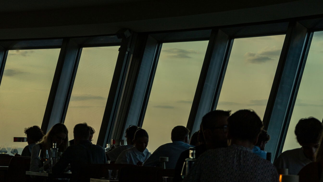 (c) Sonja Koller | 1000things Stimmungsvolle Szene beim Sonnenuntergang über Berlin: Gäste sitzen in einem Restaurant mit großen Panoramafenstern und genießen den Ausblick auf den Himmel in warmem Abendlicht. Silhouetten der Menschen heben sich gegen den goldfarbenen Horizont ab – perfekter Moment für ein romantisches Dinner mit Blick auf den Sonnenuntergang in Berlin