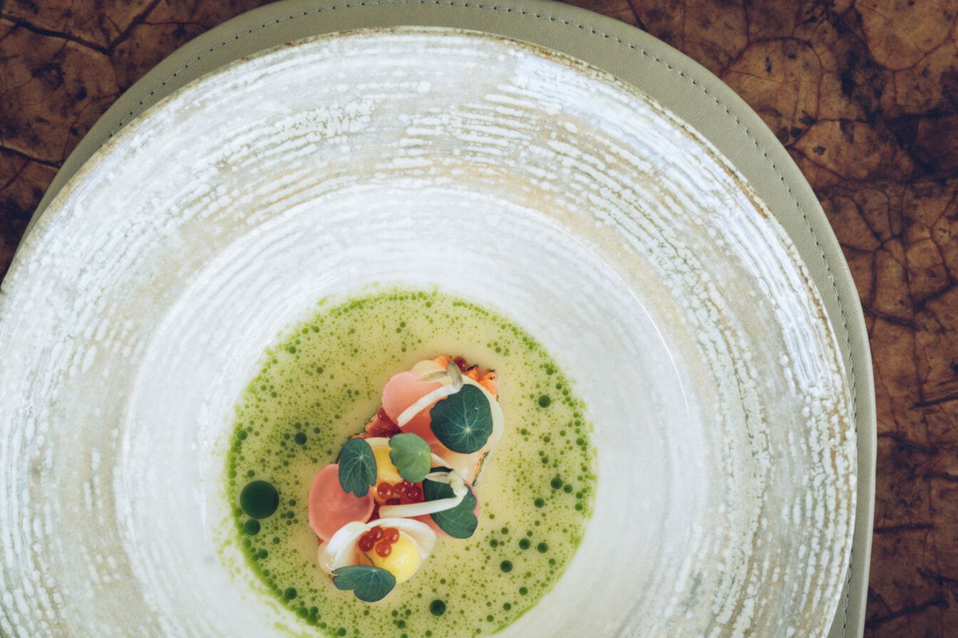 Kunstvoll angerichtetes Fine-Dining-Gericht im Restaurant The Glass Garden: zarte Komposition aus Gemüseperlen, Kräuteröl, Kaviar und Blüten auf cremiger Sauce – serviert in elegantem Keramikteller auf edlem Naturleder-Untersetzer.