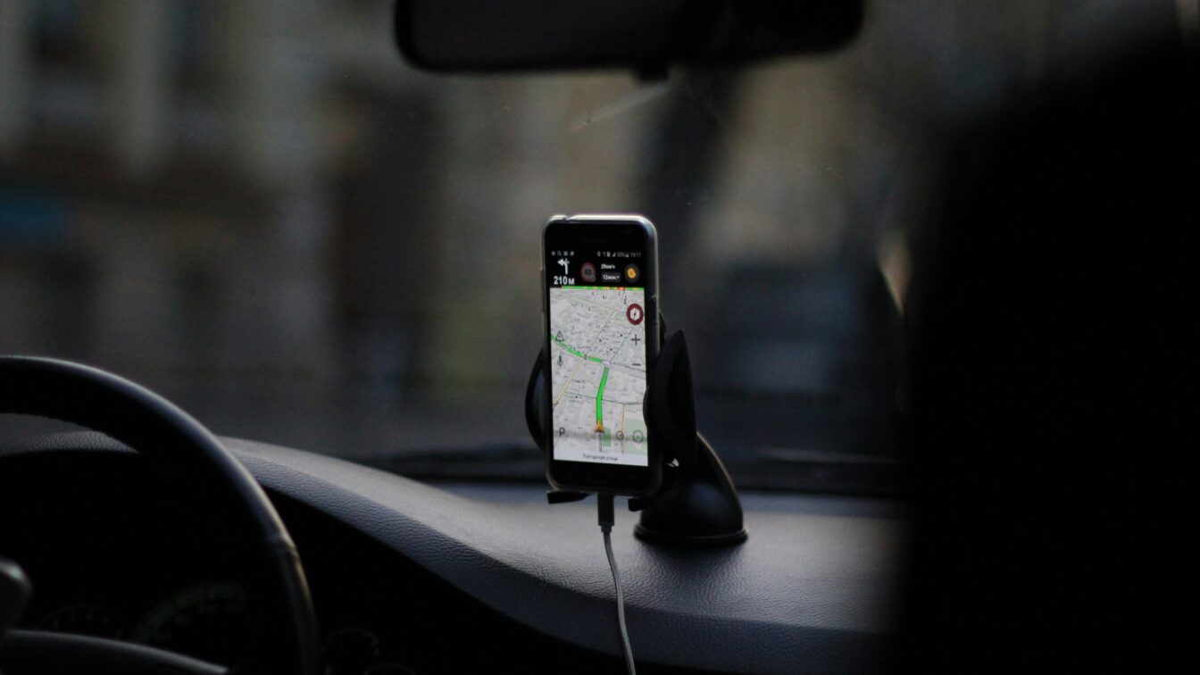 (c) Eugene Chystiakov | Unsplash Ein Smartphone in einer Halterung auf dem Armaturenbrett eines Autos zeigt eine Navigations-App mit einer geplanten Route an. Das Auto befindet sich vermutlich im Stadtverkehr, Blickrichtung durch die Frontscheibe