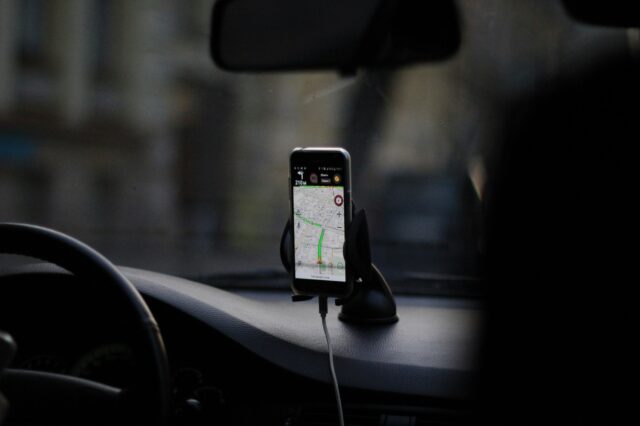 (c) Eugene Chystiakov | Unsplash Ein Smartphone in einer Halterung auf dem Armaturenbrett eines Autos zeigt eine Navigations-App mit einer geplanten Route an. Das Auto befindet sich vermutlich im Stadtverkehr, Blickrichtung durch die Frontscheibe