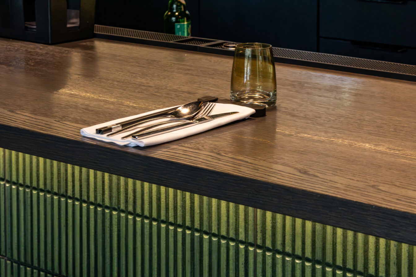 Detailaufnahme eines liebevoll gedeckten Platzes an der Bar im Vina Graz mit Besteck auf weißer Serviette und stilvollem Glas auf dunklem Holztresen.