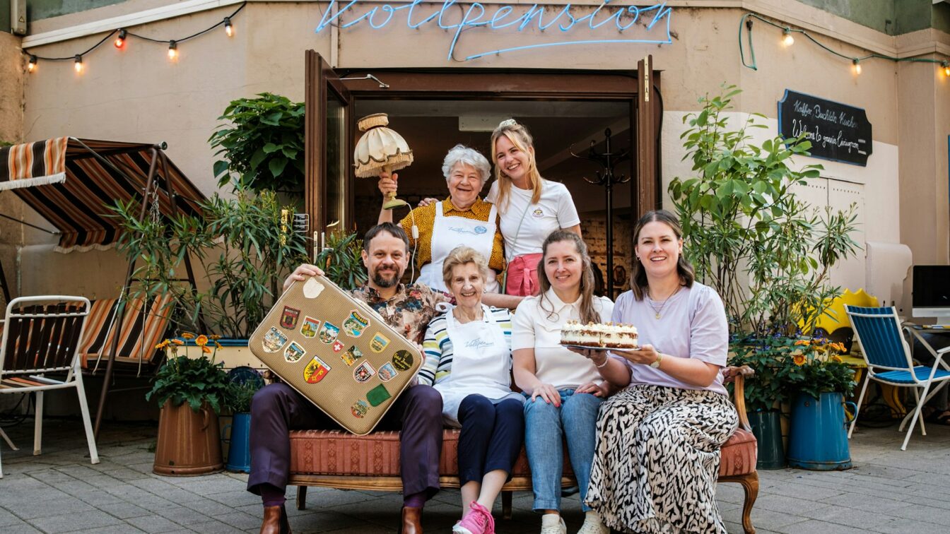 (c) Vollpension Sechs Personen vor einem Café in Wien, mit Kuchen und Koffer in der Hand.